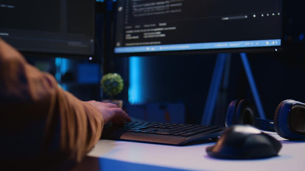 Desenvolvedor programando em ambiente escuro com múltiplos monitores, representando manutenção de código legado.