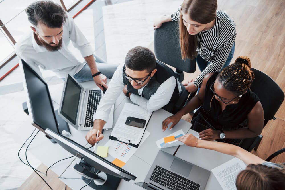 Equipe analisando dados e discutindo soluções em frente ao computador, representando colaboração em empresas de desenvolvimento de software.