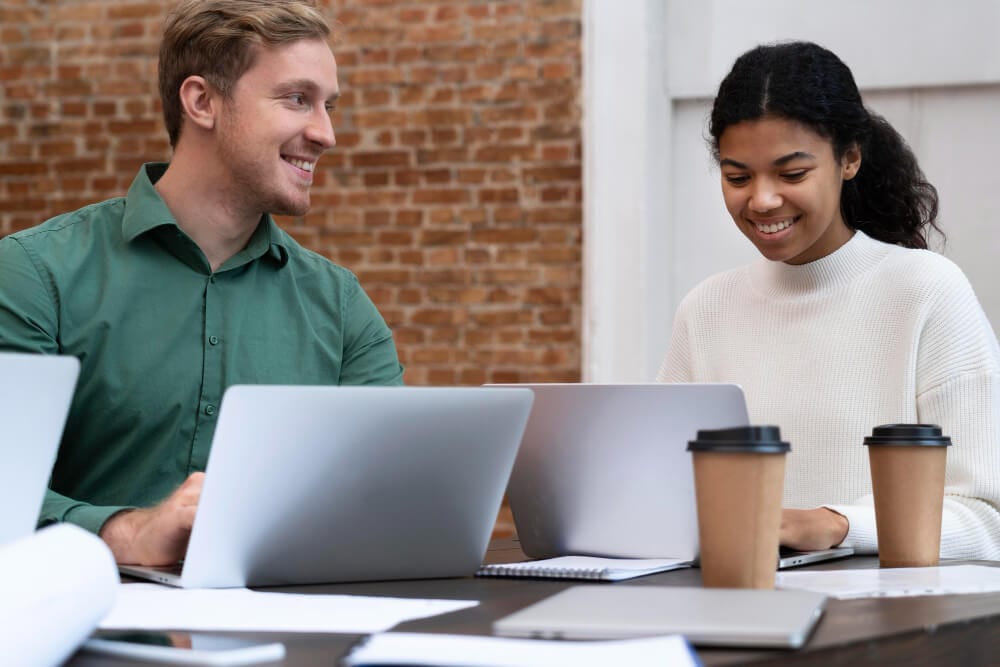 Dois colegas trabalhando juntos em notebooks, demonstrando integração de equipe e cooperação no ambiente de trabalho.