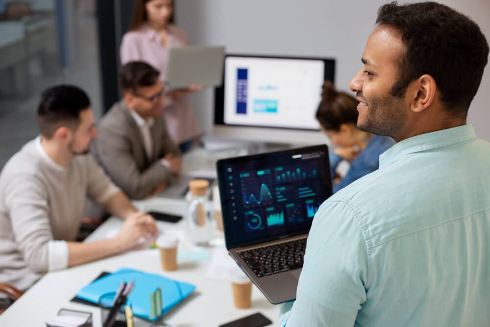 Homem segurando um notebook com gráficos de desempenho enquanto apresenta resultados para uma equipe reunida em uma sala de reunião.