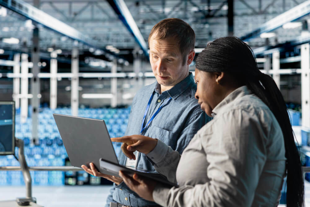 Dois profissionais analisando informações em um notebook dentro de um ambiente industrial, simbolizando gestão e desenvolvimento em uma fábrica de software.